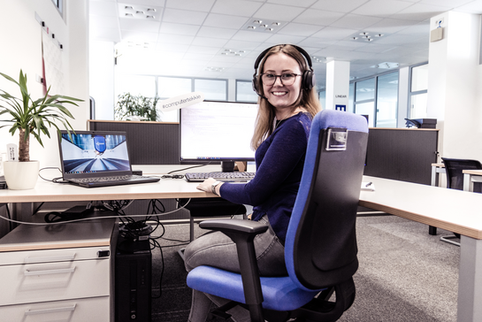 Eine junge Frau mit Headset sitzt lächelt an ihrem Schreibtisch