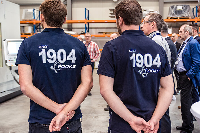 Zwei Rücken von FOOKE-Mitarbeitern. Auf ihren blauen T-Shirts steht die Jahreszahl 1904.